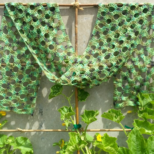 A crochet lace scarf in a variegated green yarn with plants in the background.