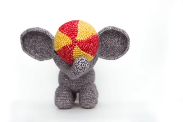 A grey crochet elephant holding a red and yellow circus ball with its trunk.