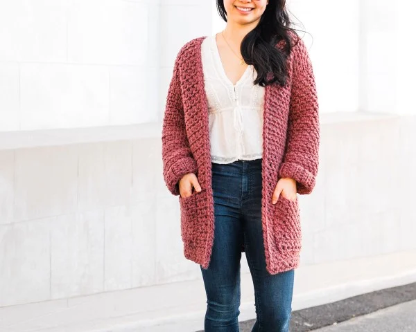 A woman wearing a rose pink coloured crochet cardigan with pockets and jeans.