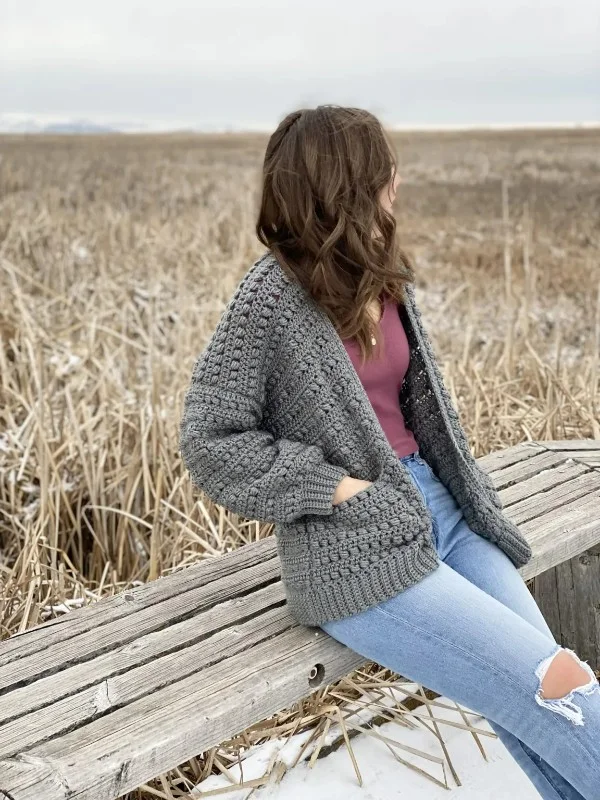 A woman sitting outdoors wearing a grey crochet cardigan with pockets.