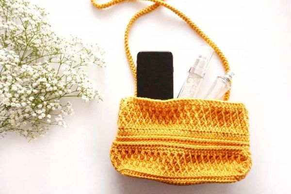 A bright yellow crochet crossbody bag with a mobile phone and a bunch of flowers.