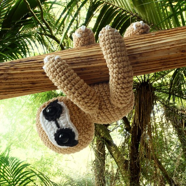 A crochet sloth hanging upside down on a tree with a tropical forest background.