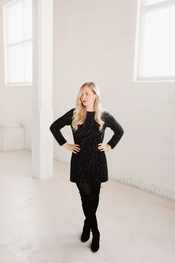 A woman wearing a black crochet sweater dress with tights and boots.