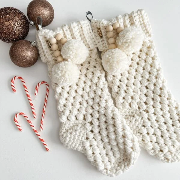 Two white crochet Christmas stockings with pompoms next to candy canes and Christmas baubles.