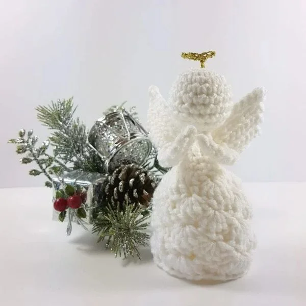 A white crochet angle with a golden halo next to a pine cone and a branch from a pine tree.