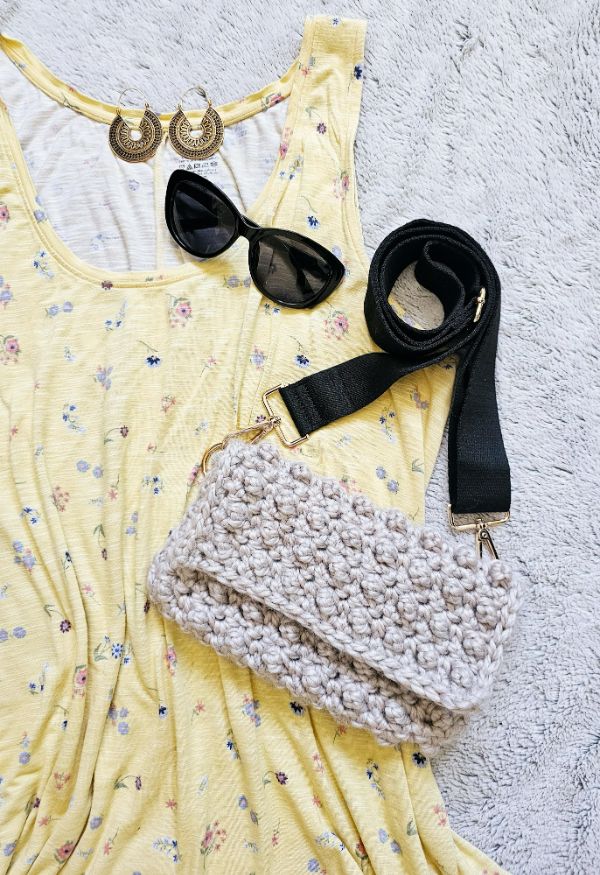 A flat lay of a summer dress, sunglasses, and a crochet crossbody bag with a black strap.