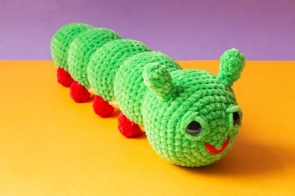 A green crochet caterpillar with red feet worked in cuddly chenille yarn.
