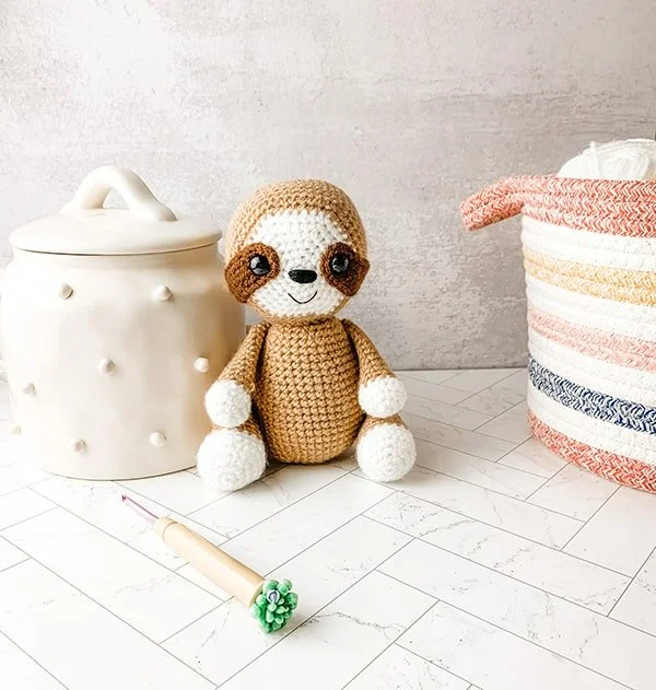 A small brown crochet sloth sitting on a bench.