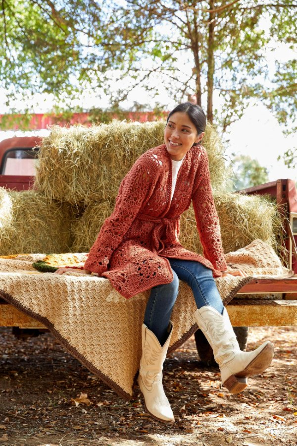 A woman sitting on the back of a truck wearing a long crochet granny square cardigan.