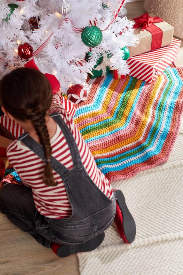 A child placing presents under a Christmas tree with a brighlty coloured, striped crochet Christmas tree skirt.