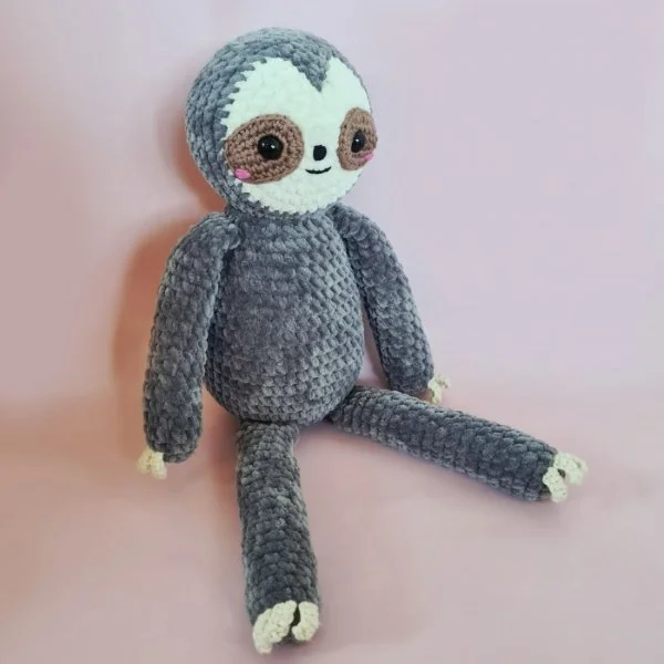 A grey crochet sloth toy on a pink background.