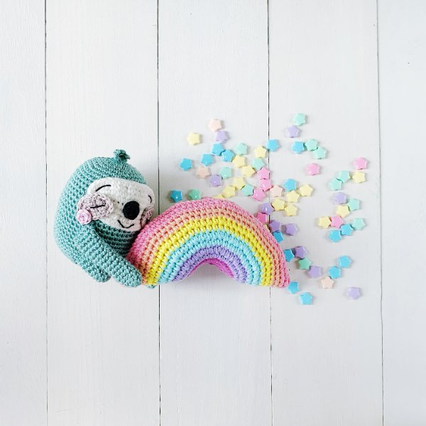 A cute blue crochet sloth cuddling a rainbow.