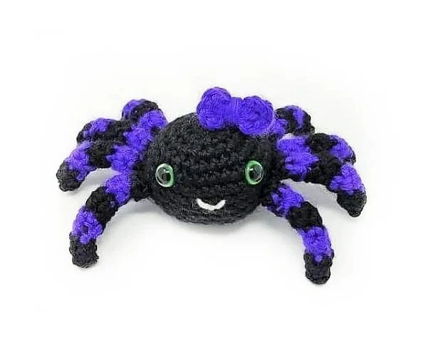A purple and black crochet spider with a purple bow and green eyes.
