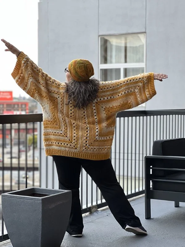 Back view of a woman wearing a yellow toned crochet hexagon cardigan with black pants.