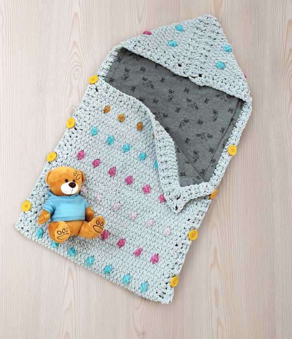 A crochet sleeping bag for babies pictured with a little teddy bear.