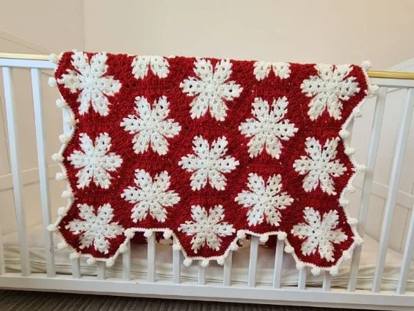 A red and white crochet snowflake Christmas blanket hanging over the side of a crib.
