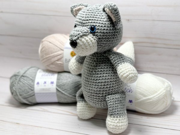 A crocheted wolf softie toy standing next to balls of yarn.