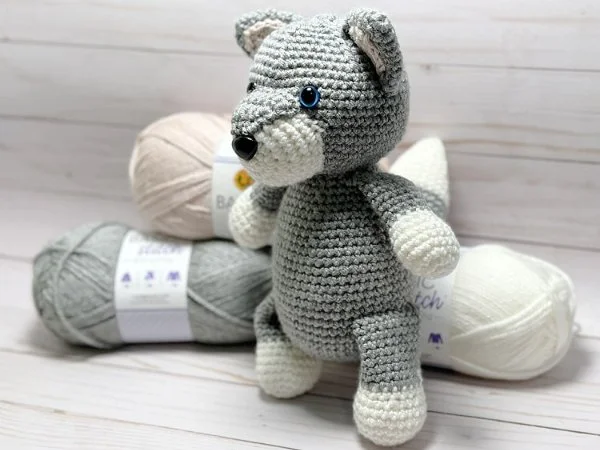 A crocheted wolf softie toy standing next to balls of yarn.