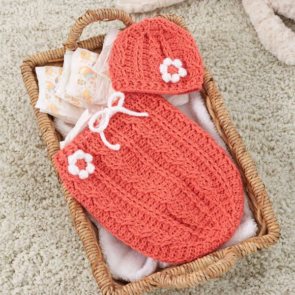 A cable crochet baby cocoon and matching hat laid out in a basket.