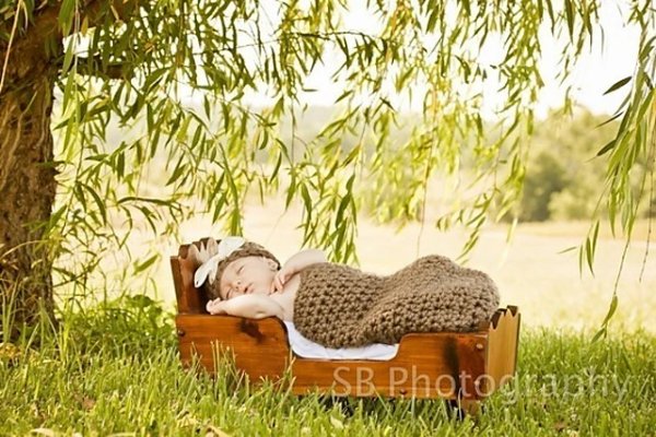 A baby in a brown crochet baby cooon sleeping in a wooden cradle in a garden.