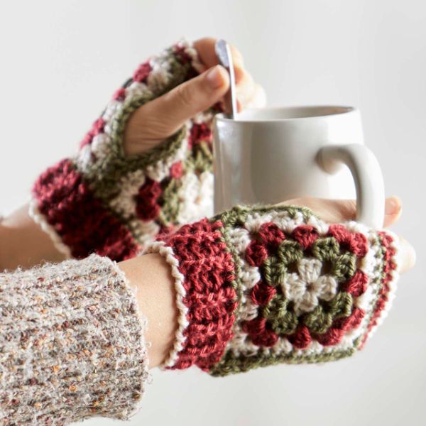 A close up image of a woman holding a cup of coffee while wearing granny square fingerless gloves.