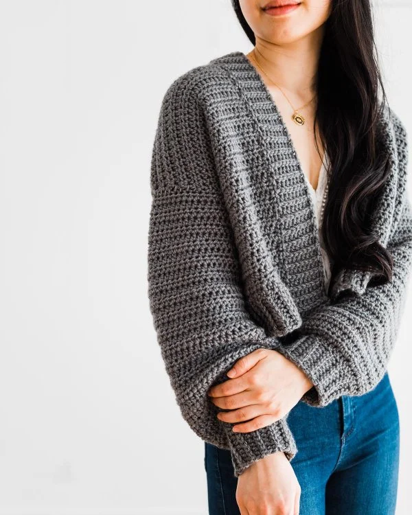 A woman aearing a grey, oversized, cropped crochet cardigan.