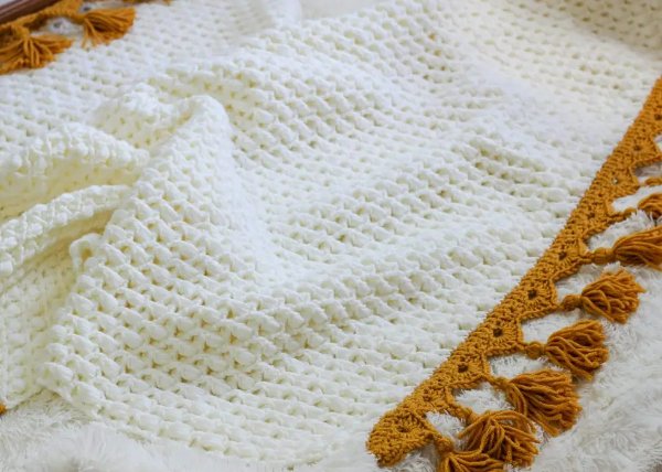 Close-up image of a white crochet blanket with golden yellow edging and tassels.