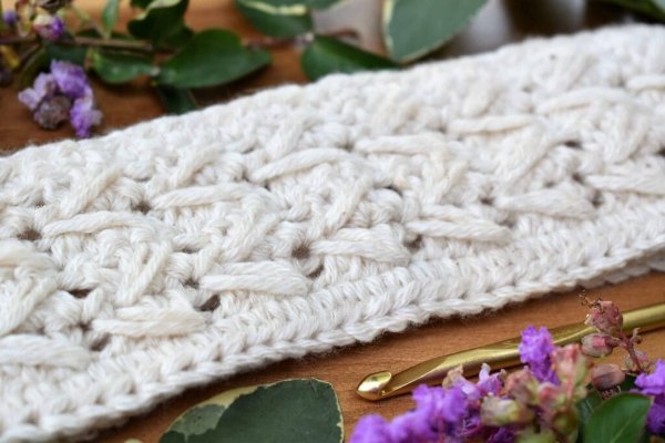 A closeup image of a white crochet headband featuring a cble stitch design.