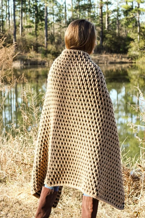 A woman outdoors with a chunky crochet blanket wrapped around her.