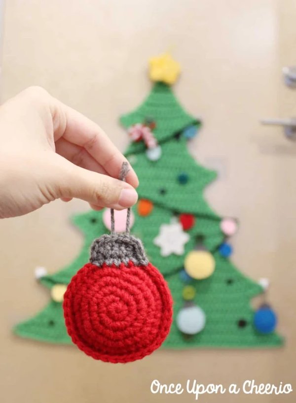 A simple red crochet bauble, with a crocheted Christmas tree in the background.