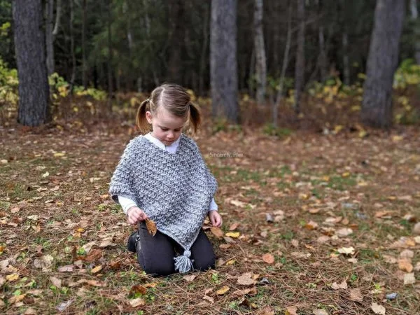 A child surrounded by fall leaves wearing a grey crochet poncho with tassels