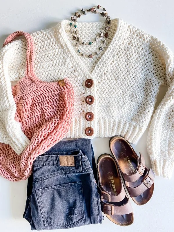 A flat lay image of a cropped crochet cardigan with jeans and sandals.