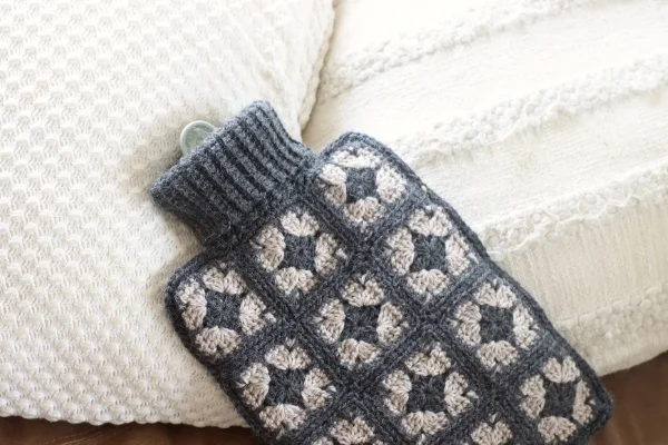 A granny square hot water bottle cover with white cushions.