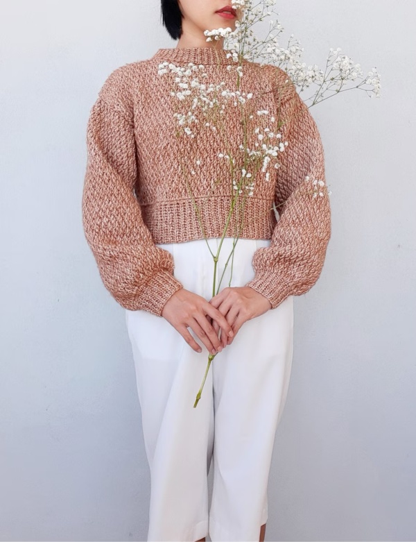 A woan wearing an oversized crochet cropped sweater with white pants.