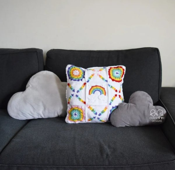 A white crochet granny square pillow with rainbow coloured accents on a couch.