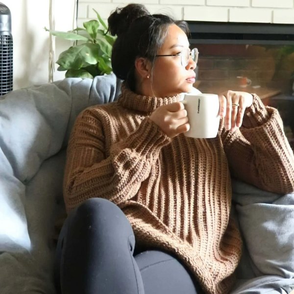 A woman drinking coffee on a sofa, wearing a ribbed crochet turtleneck sweater.