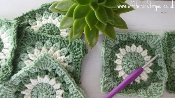 A pile of green crochet granny squares iwth a crochet hook and  a plant in the background.