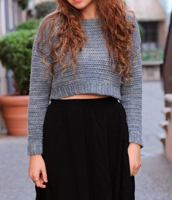 A woman weaaring a grey cropped crochet sweater with a black skirt.