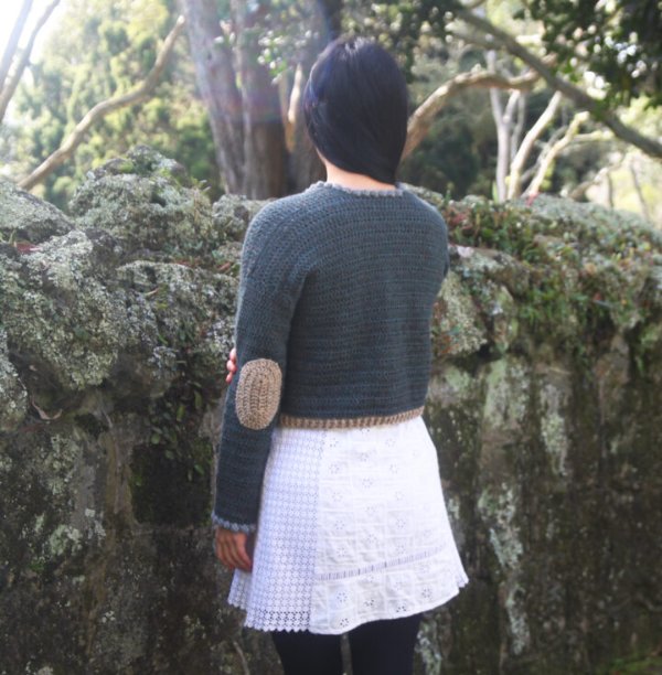 Back view of a woman wearing a grey, croppeed crochet cardigan with elbow patches.