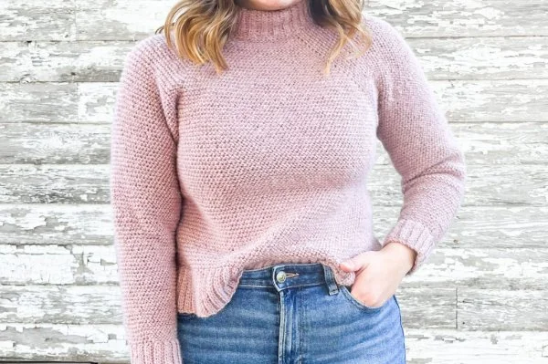 A closeup of a woman weraing jeans and a pink, turtleneck, raglan crochet sweater.