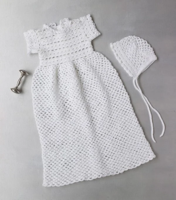A flat lay image of a short-sleeved crochet christening gown and a bonnet.