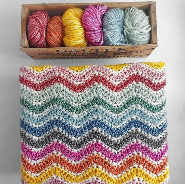 A folded rainbow crochet baby blanket next to a basket of yarn.