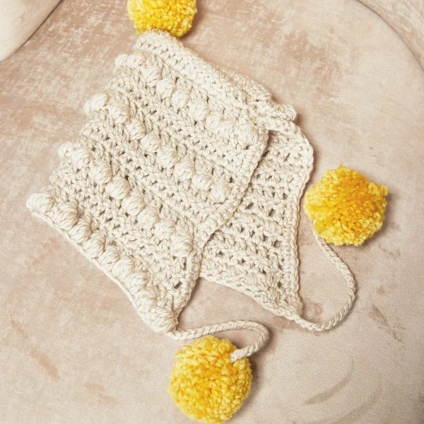 A white crochet baby bonnet with yellow pompoms.