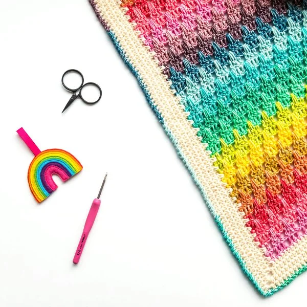 A rainbow crochet baby blanket with scissors and a crochet hook.