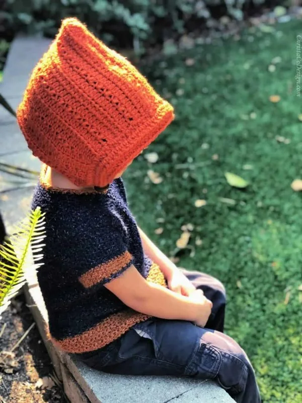 A toddler in a garden wearing an orange crochet bonnet.
