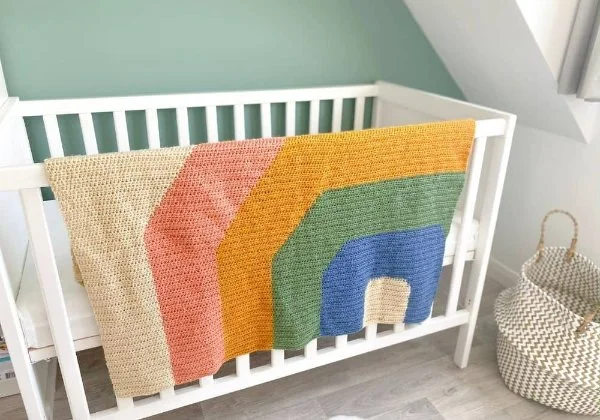 A rainbow crochet baby blanket hanging over a crib.