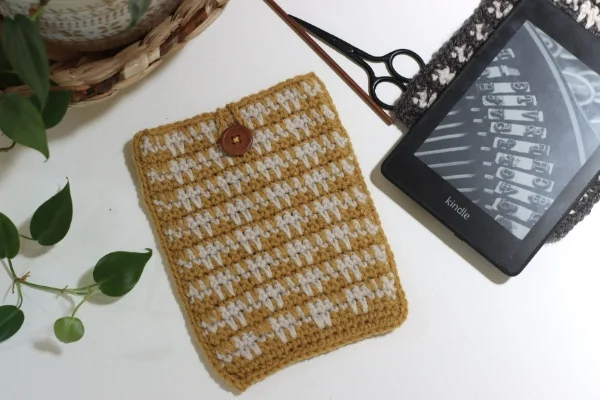 A yellow and white crochet kindle sleeve with a kindle and crochet supplies.