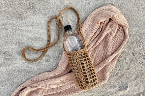 A crochet bottle bag on a towel at the beach.