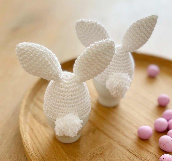 Two white crochet egg cozies with bunny ears and fluffy bunny tails.