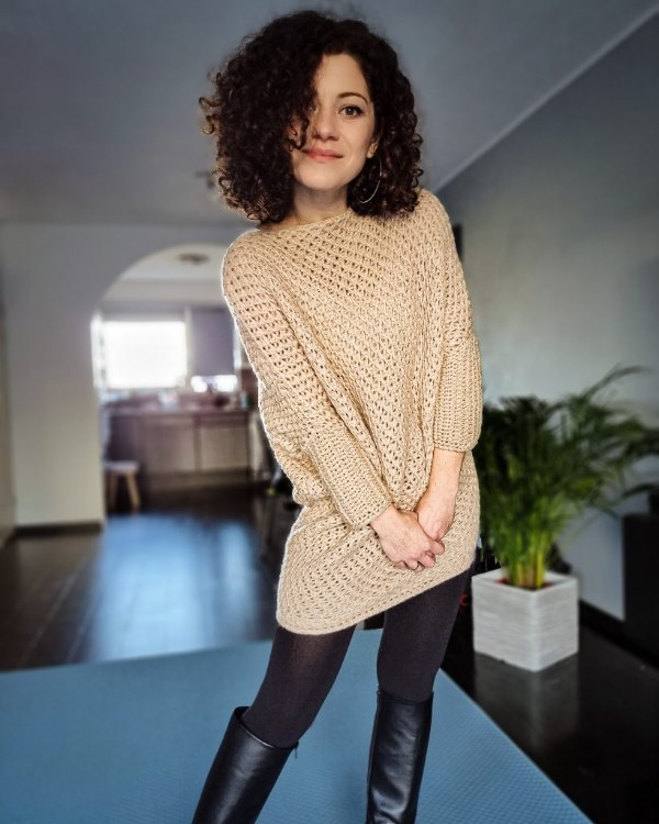 A woman wearing a straw coloured crochet sweater dress with black leggings and boots.